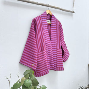 Purple striped garment hanging on a wooden hanger against a white wall with green plants at the bottom.