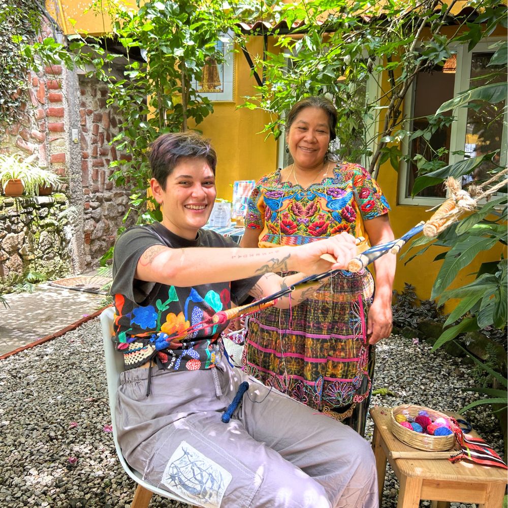 Two people in a garden setting, one holding a colorful loom, with a yellow building in the background.