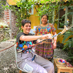 Two people in a garden setting, one holding a colorful loom, with a yellow building in the background.