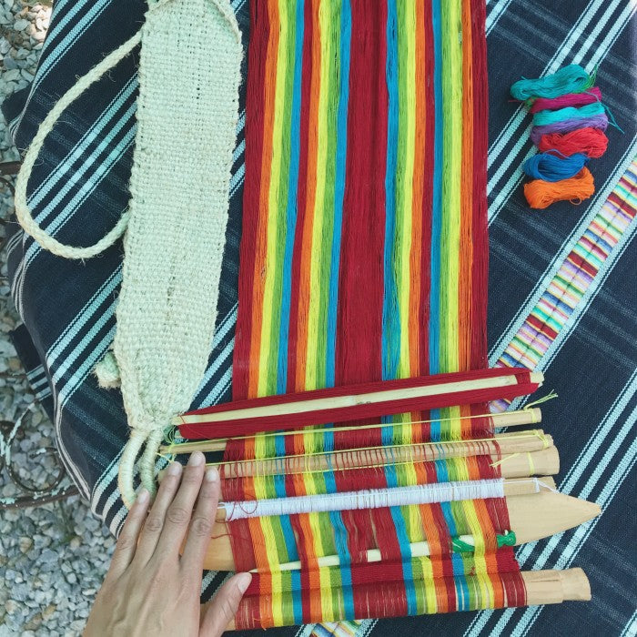 A colorful brocade weaving project on a backstrap loom with various tools and materials laid out, including extra rods, a wooden needle, and multiple threads.
