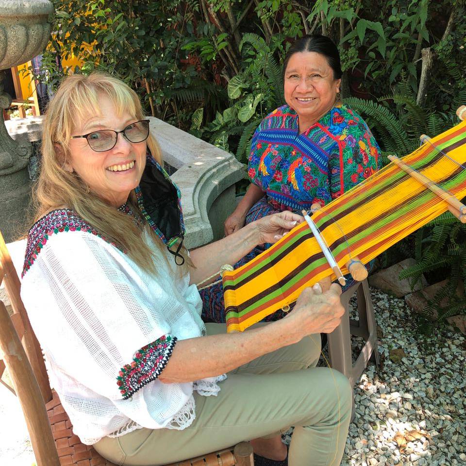 Two women smiling with a colorful loom in a lush garden